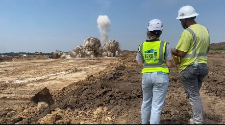 Two people in hard hats watch a blast from a distance