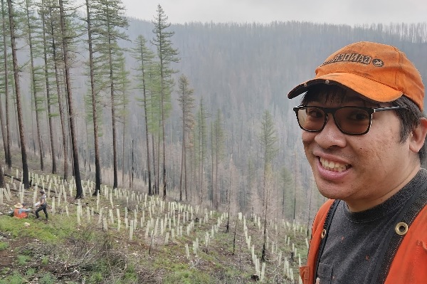 !Matthew Dang stands on a mountainside with tree saplings.